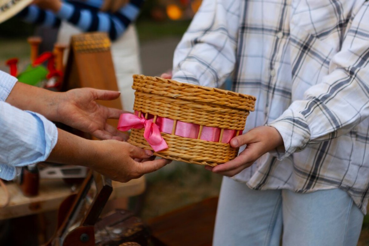 Adult hands giving a woven basket with a pink ribbon to a child at an outdoor market.