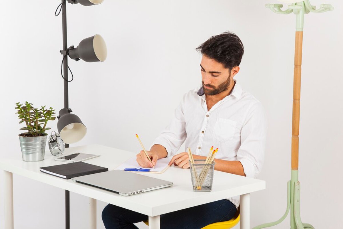 A focused man writes in a notebook at a tidy desk with a laptop and plant.