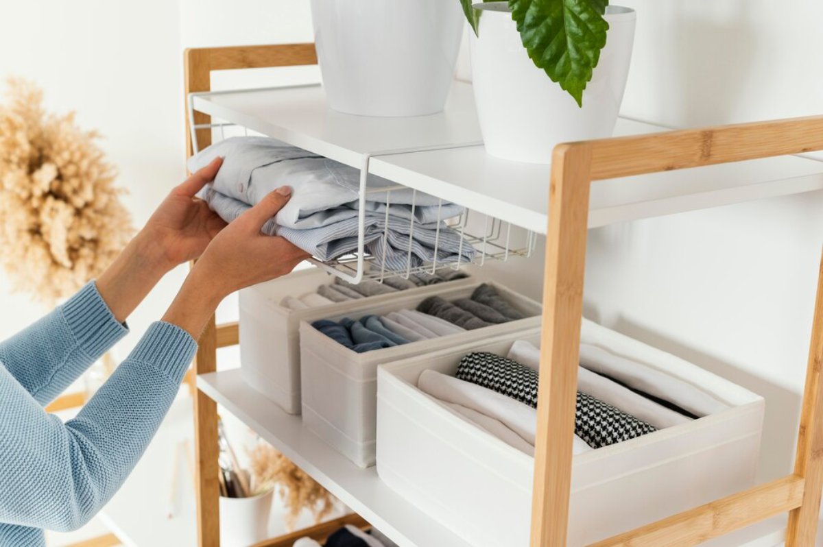 Person organizing neatly folded clothes into a modern, open shelved closet with storage boxes and decorative plants.