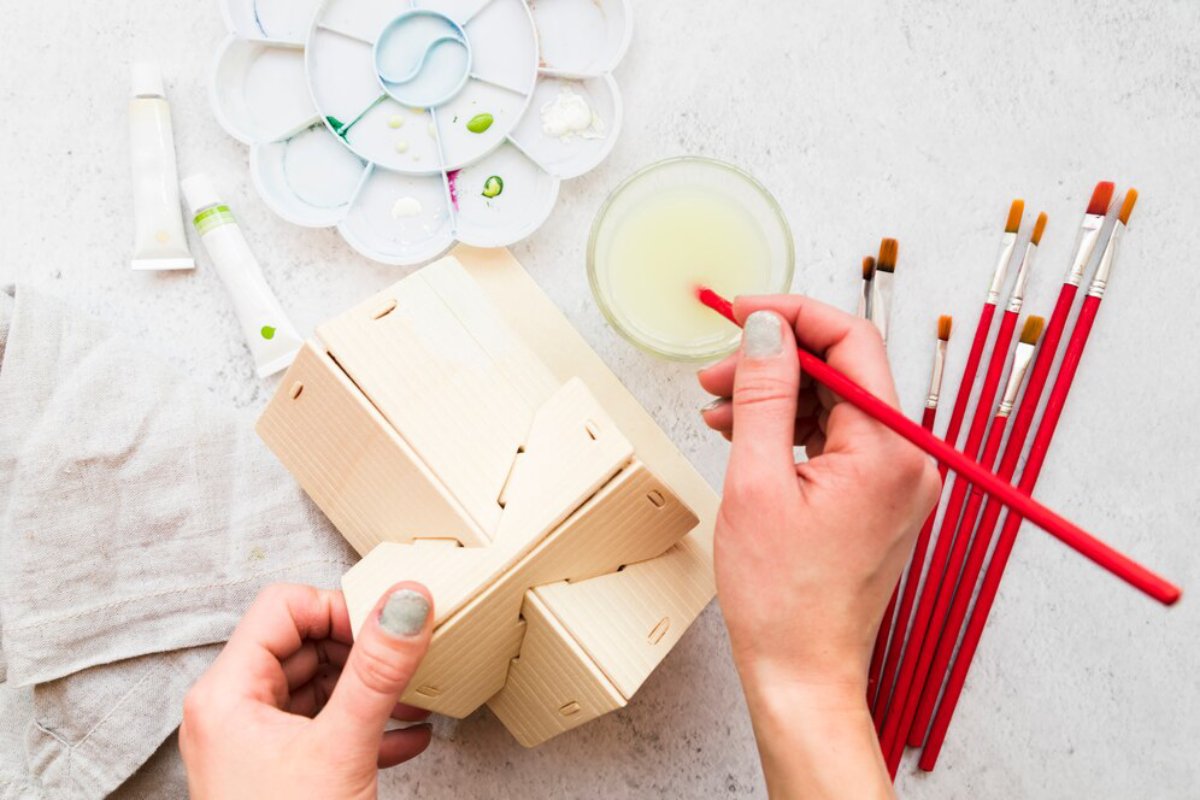 Hands painting a wooden birdhouse with white paint, surrounded by brushes and paint tubes on a light background.