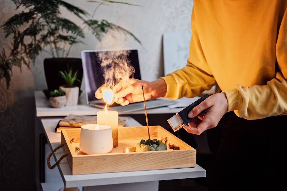 Person lighting a candle on a wooden tray with plants and a laptop in the background.