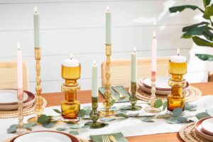 Elegant dining table setup with lit candles in decorative holders, greenery, and stylish dishes on a white background.