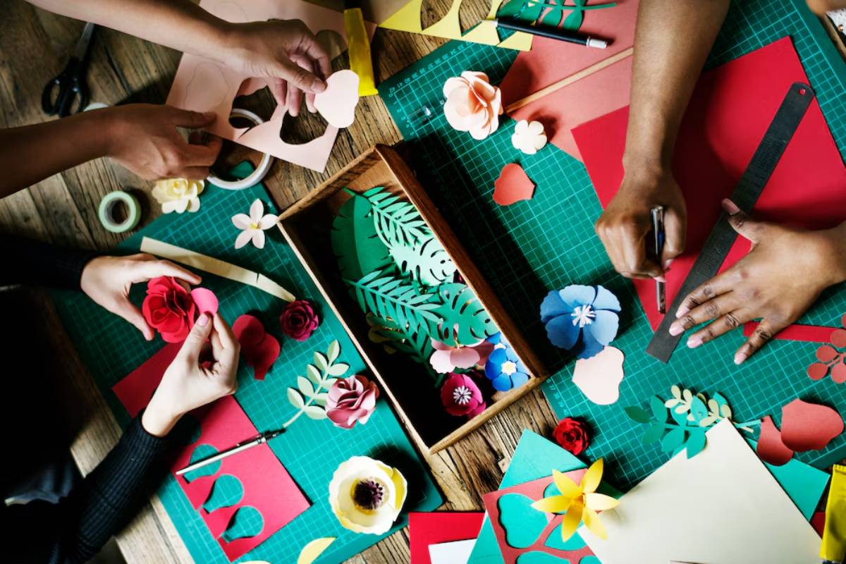 Two people crafting with colorful paper, making floral designs on a wooden table surrounded by crafting tools and materials.