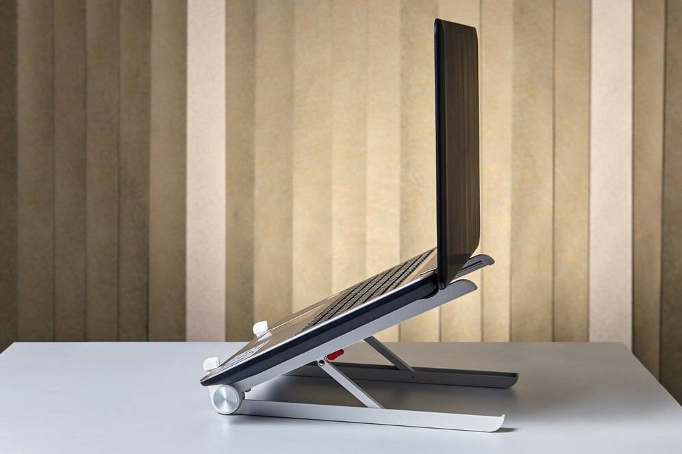 A modern laptop stand on a table with a closed laptop on it, against a textured beige background.