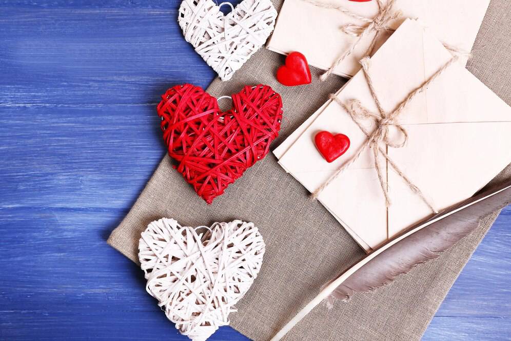 Decorative wicker and red heart shapes next to wrapped gifts on a blue wooden background.