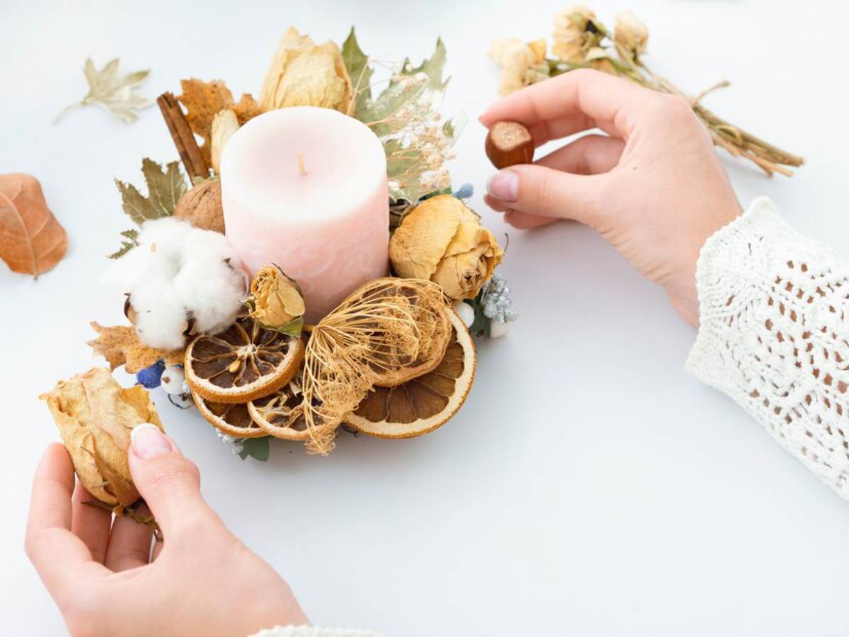 Hands arranging a decorative autumn centerpiece with dried oranges, flowers, and a pink candle on a white background.