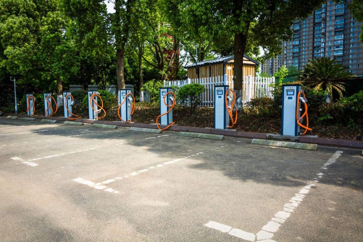 A row of electric vehicle charging stations in an outdoor parking lot with trees and a building in the background.