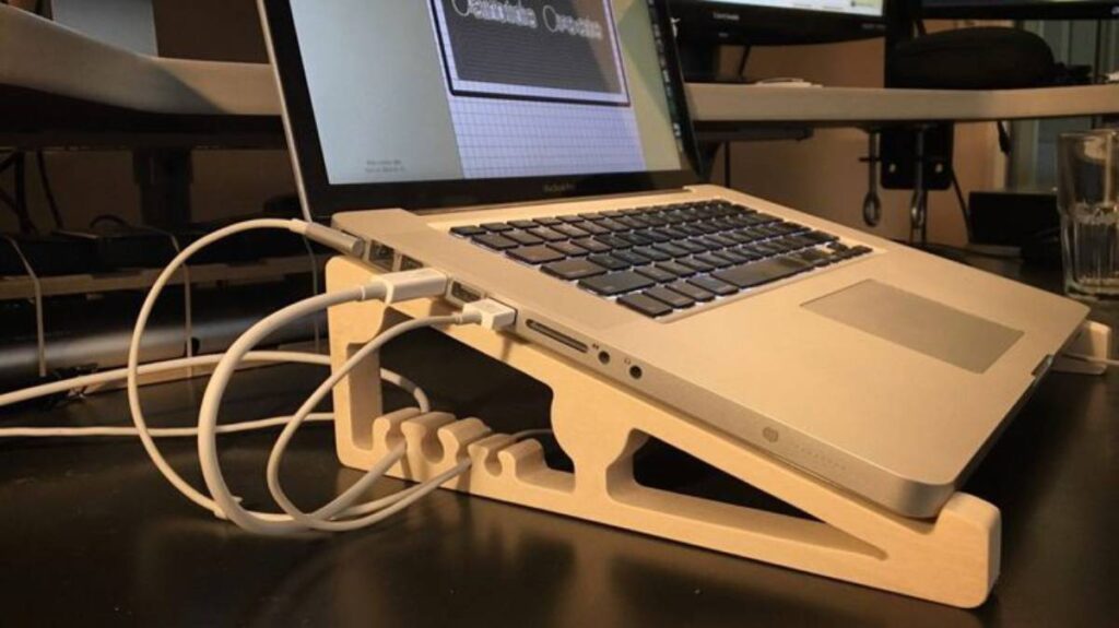 A laptop on a wooden stand connected to multiple cables, with a black monitor in the background.
