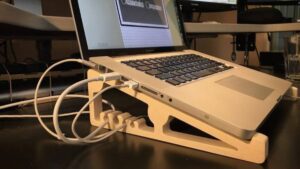 A laptop on a wooden stand connected to multiple cables, with a black monitor in the background.