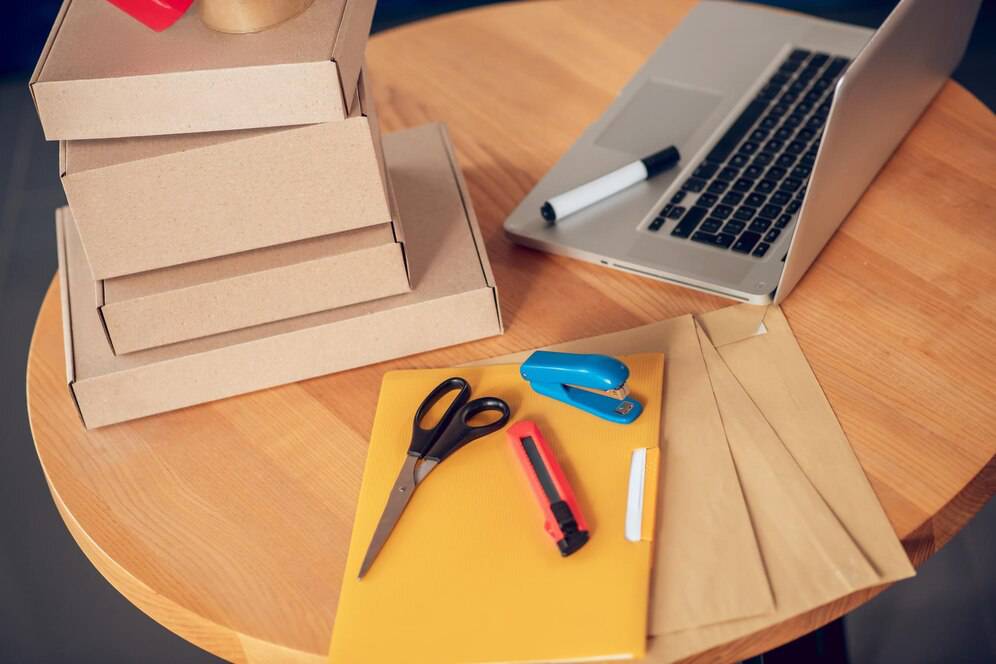 A workspace with stacked cardboard boxes, a laptop, and packing supplies on a wooden table.