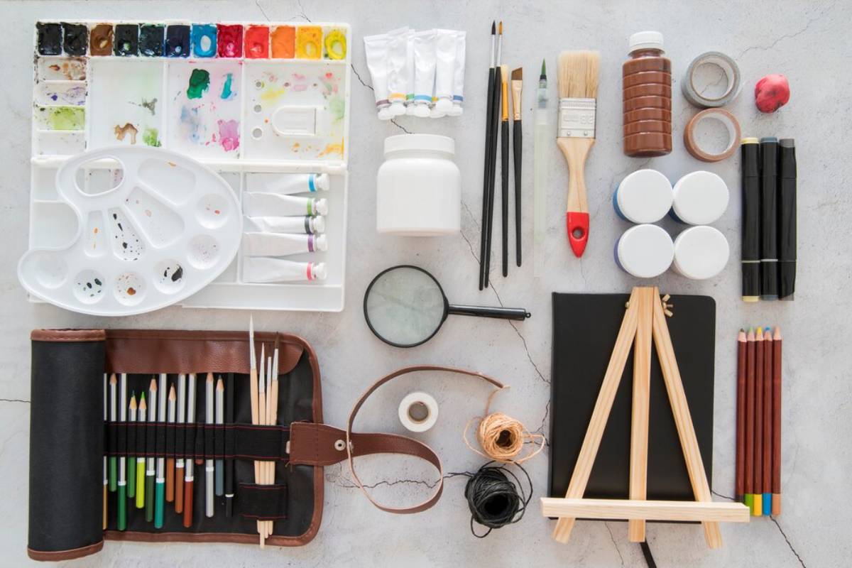 Assorted art supplies including watercolors, brushes, colored pencils, and a small easel laid out on a gray surface.