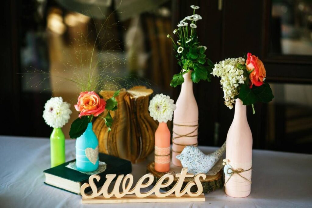 Decorative table display featuring upcycled bottles as vases with colorful flowers and a wooden "sweets" sign.