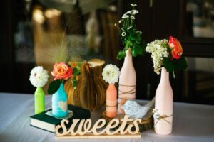 Decorative table display featuring upcycled bottles as vases with colorful flowers and a wooden "sweets" sign.