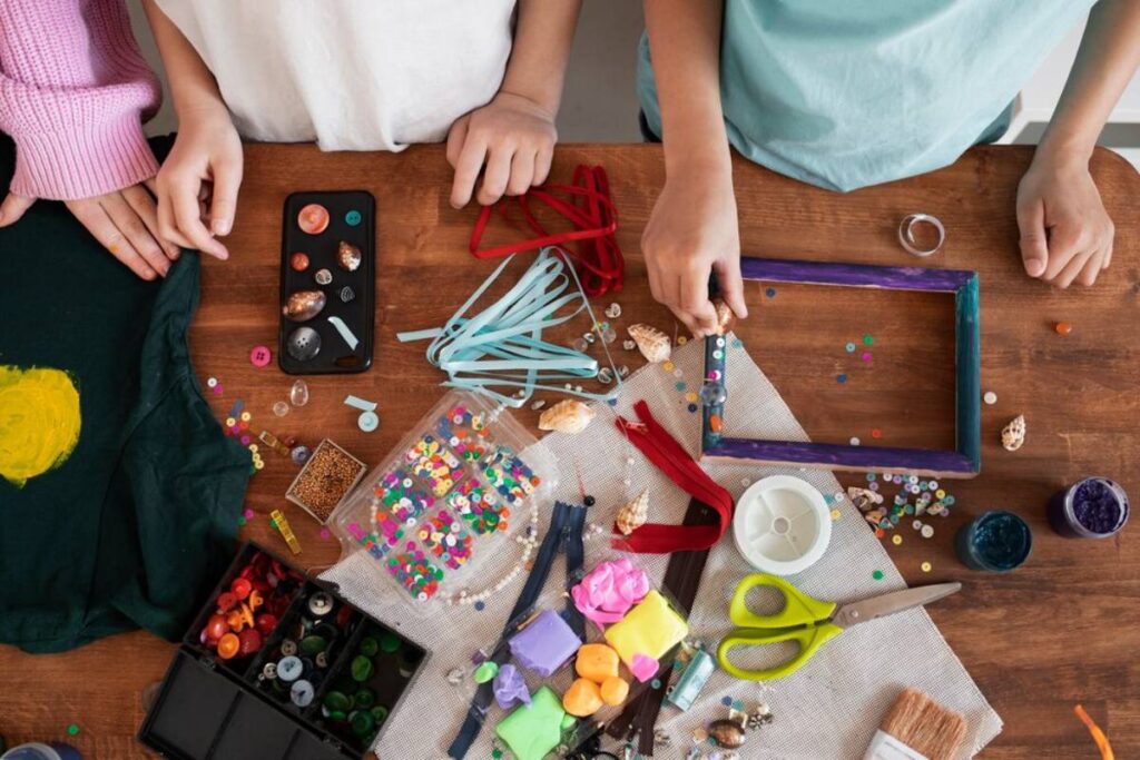 Overhead view of children's hands crafting with beads, buttons, paints, and fabric on a wooden table.