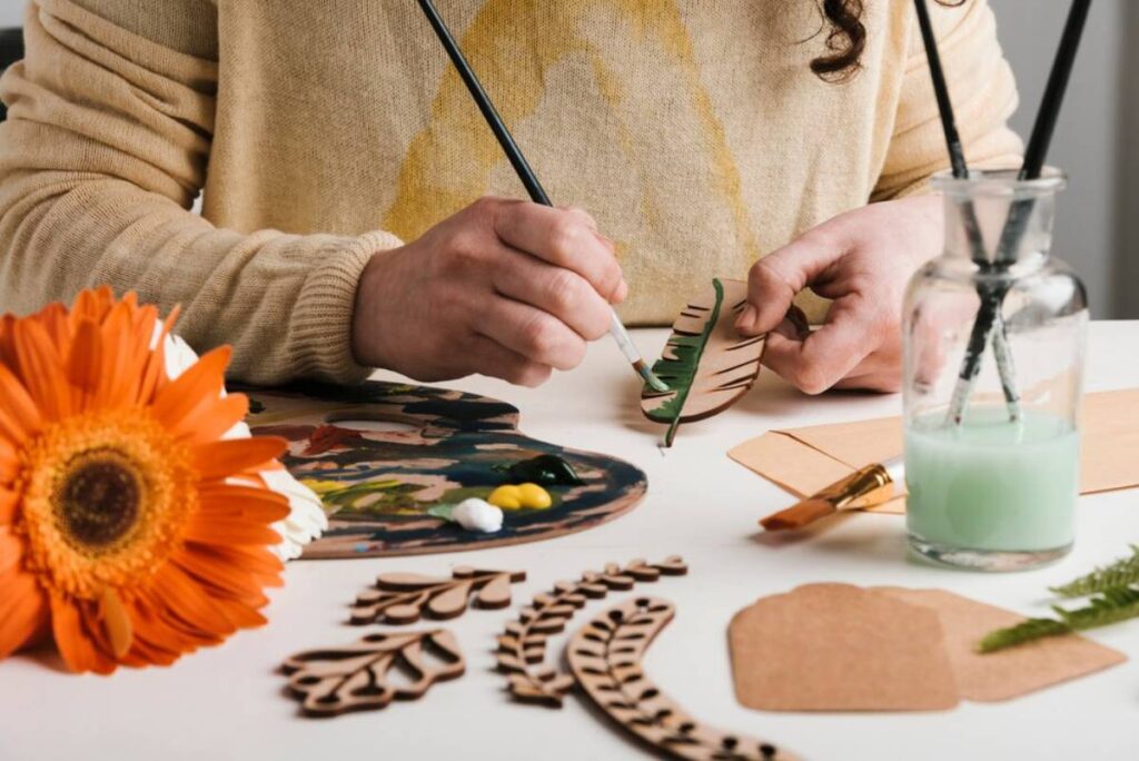 Person painting a wooden leaf cutout, surrounded by art supplies and a bright orange flower on the table.