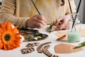 Person painting a wooden leaf cutout, surrounded by art supplies and a bright orange flower on the table.