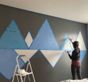 Person painting blue mountain shapes on a dark grey wall, standing on a ladder.