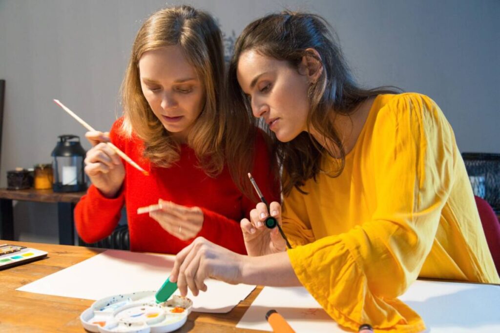 Two women concentrating on painting with watercolors at a table in a softly lit room.