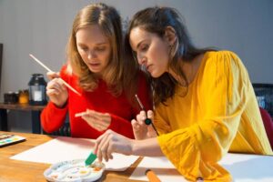 Two women concentrating on painting with watercolors at a table in a softly lit room.