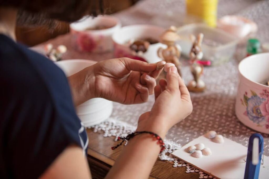 A person engaging in a craft, handling small shells and decorative items on a table.