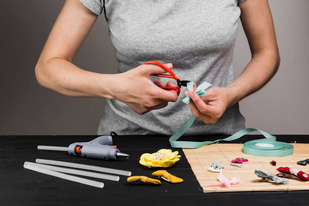 A woman using scissors to cut ribbon at a crafting table with various craft tools and materials.