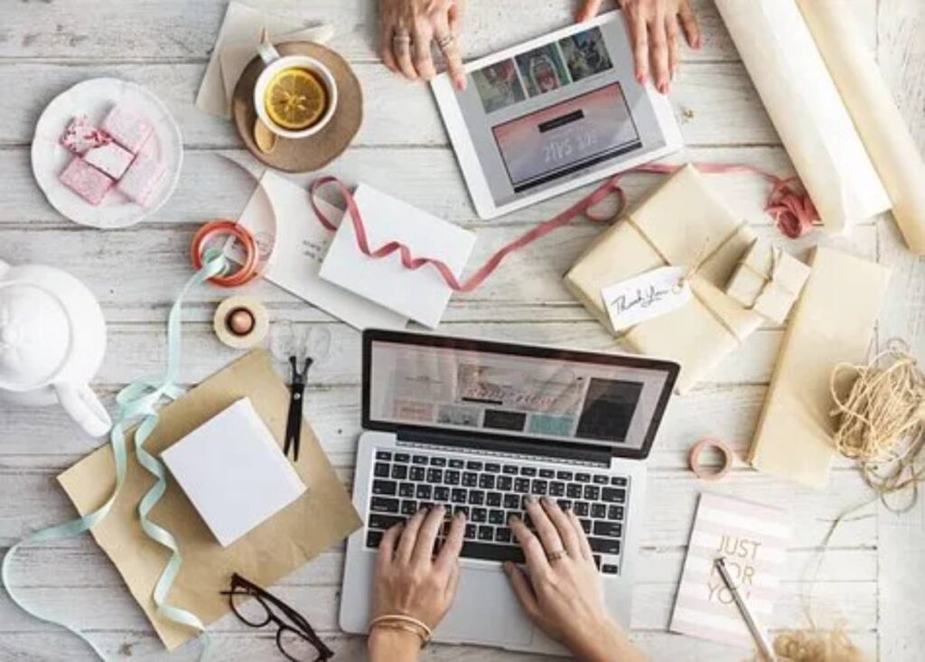 Overhead view of a person's hands working on a laptop surrounded by gift wrapping materials and a cup of tea.