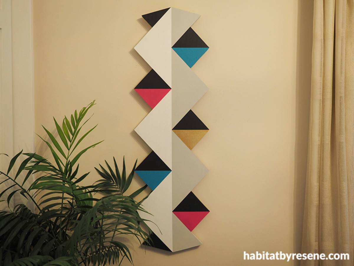 A modern, geometric wall art piece in white, black, and bright colors next to a potted plant and cream curtains.