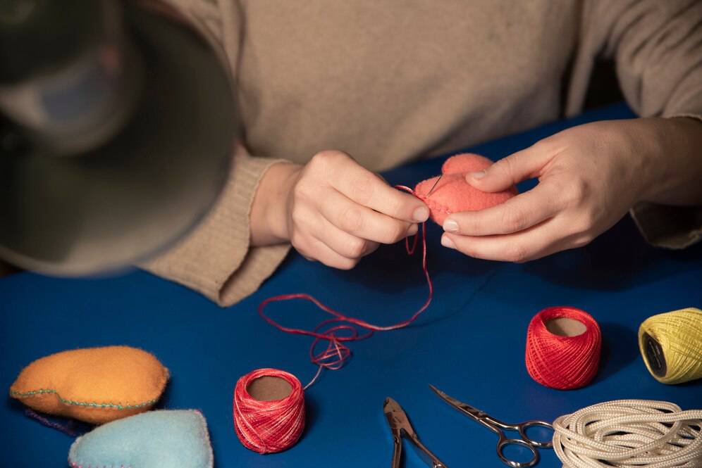 Person sewing a red heart-shaped fabric with needle and thread on a blue table, surrounded by crafting supplies.