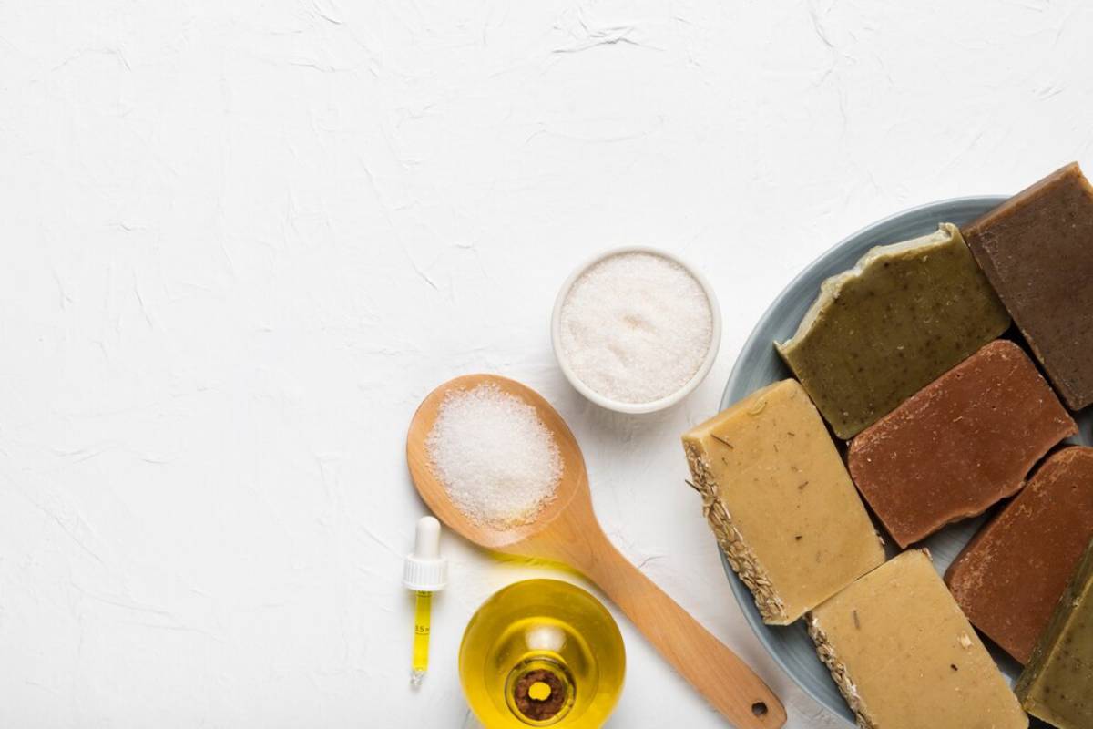 Handmade soap bars on a plate with oil dropper, salt on wooden spoons, and white textured background.