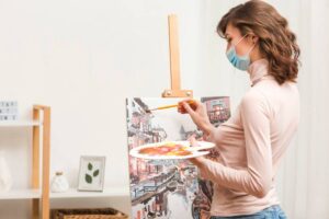 Woman in a mask painting a colorful street scene on canvas in a bright room.