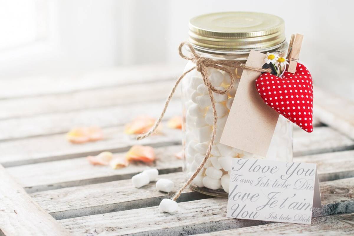 A glass jar tied with twine and a heart-shaped decoration, filled with white pebbles, on a rustic wooden background.