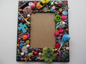 A picture frame decorated with colorful assorted buttons, toys, and trinkets surrounding a blank rectangular space.