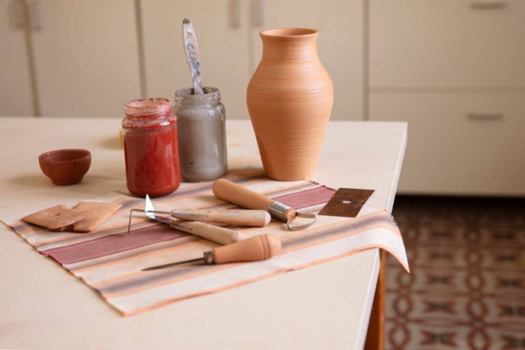Pottery tools, a clay vase, and jars of glaze on a striped cloth on a table.