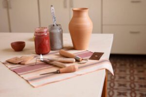 Pottery tools, a clay vase, and jars of glaze on a striped cloth on a table.