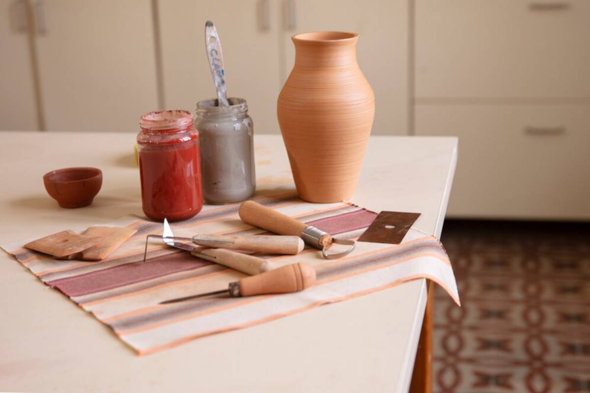 Pottery tools, a clay vase, and jars of glaze on a striped cloth on a table.