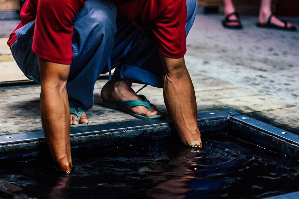 Person squats and dips feet in shallow water basin, wearing flip-flops and casual clothing.