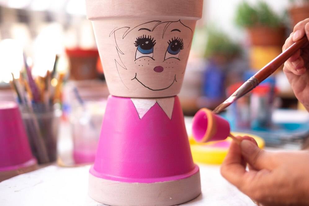 A person painting a smiling face on a terracotta pot shaped like a cartoon character, in a colorful craft environment.