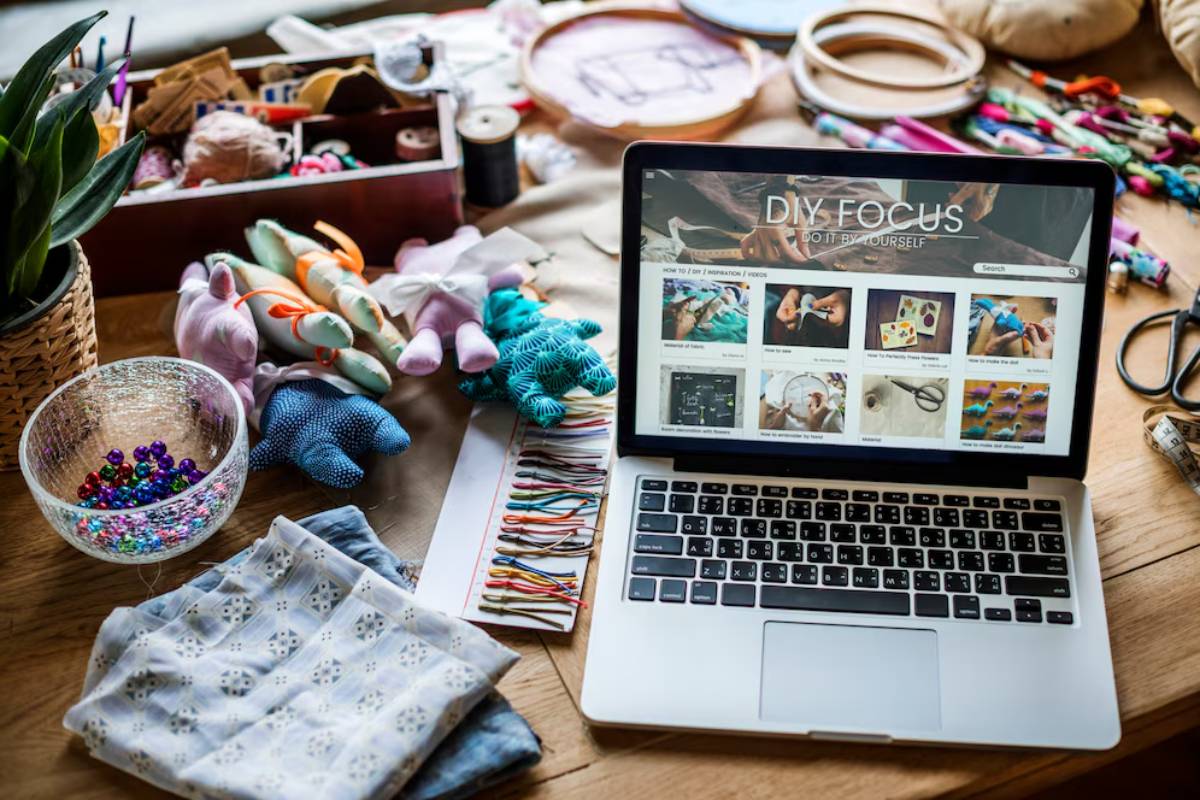 A laptop displaying a DIY craft website surrounded by handmade toys, sewing materials, and fabric on a wooden table.