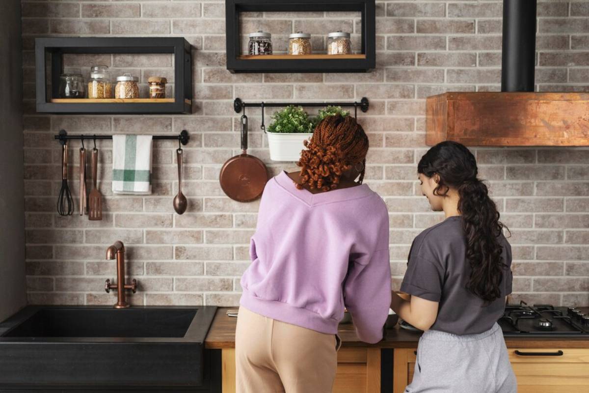 Two women cooking together in a modern kitchen with brick wall and hanging utensils.