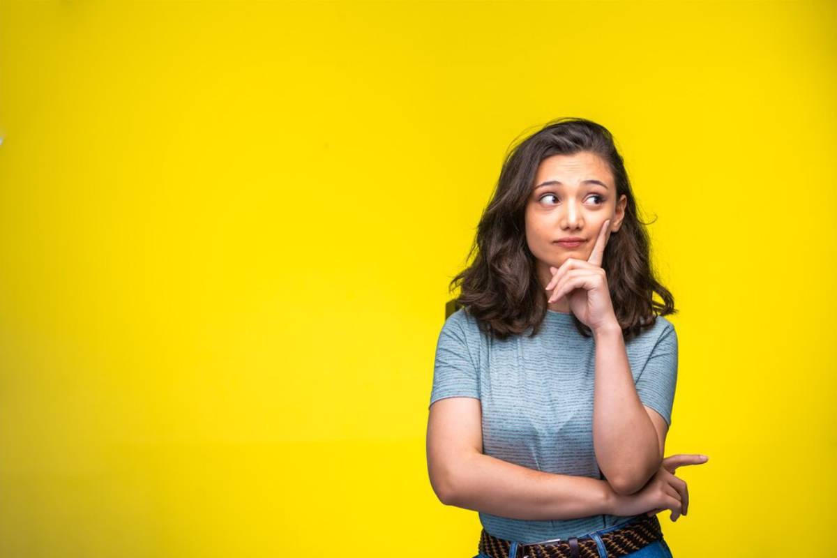Young woman pondering with a finger on her cheek, standing against a bright yellow background.