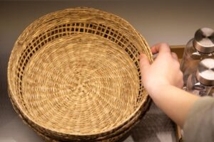 A hand placing a woven wicker basket on a shelf next to metal containers.