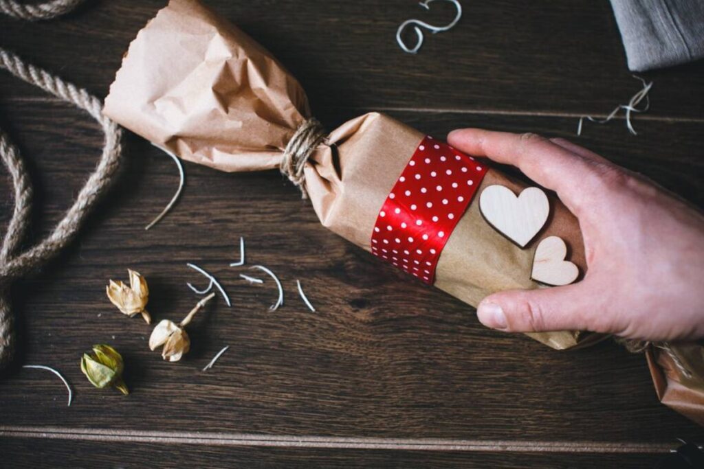 Hand wrapping a gift with brown paper, red polka-dot ribbon, and wooden heart decorations on a wooden table.