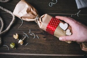 Hand wrapping a gift with brown paper, red polka-dot ribbon, and wooden heart decorations on a wooden table.
