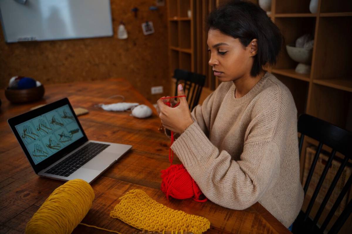 Woman knitting at a table with a laptop displaying crochet patterns, surrounded by yarn.