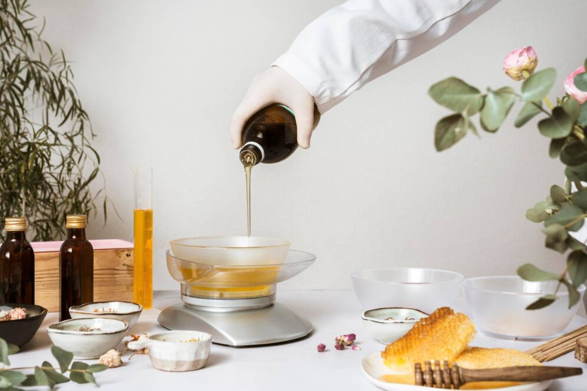 A person pouring oil into a bowl on a scale, surrounded by ingredients and cosmetic bottles in a serene setting.