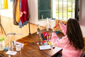 A young girl crafts at a wooden table near a window, with craft supplies scattered around.
